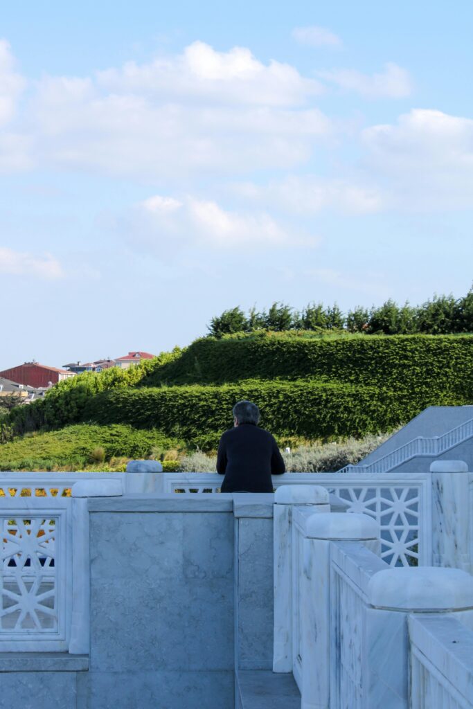 A man stands by a marble balcony overlooking a lush garden, enjoying the serene summer surroundings.