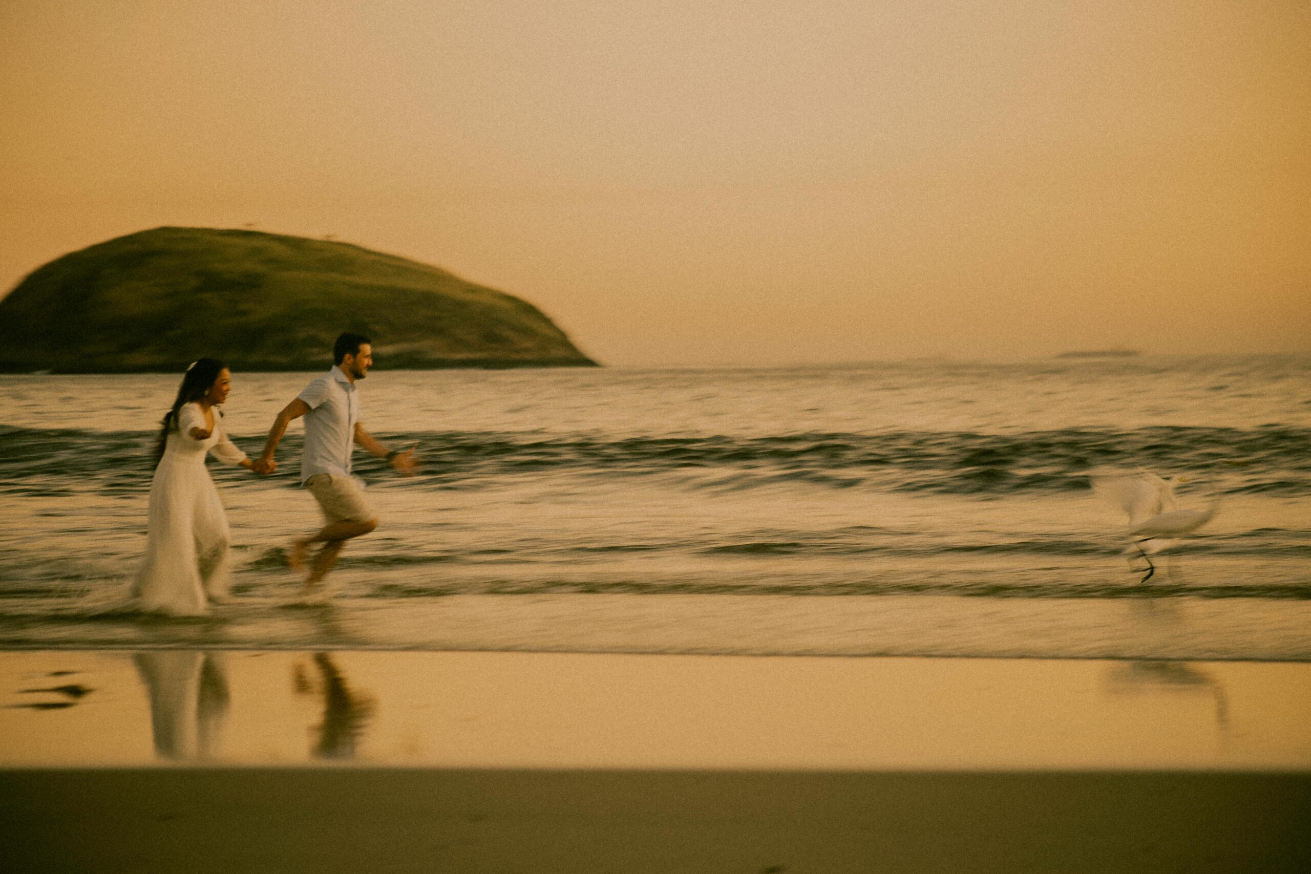 A couple runs joyfully on a beach at sunset with a scenic view of Rio de Janeiro.
